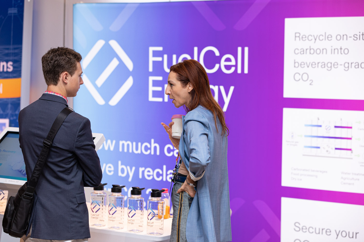 A man in a navy blazer and a woman in a blue jacket stand in front of a purple FuelCell Energy display about recycling carbon into beverage-grade CO2.