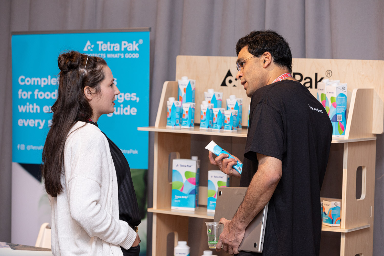 A woman in a white cardigan and a man in a black T-shirt look at cartons on a wooden shelf in front of a Tetra Pak banner.