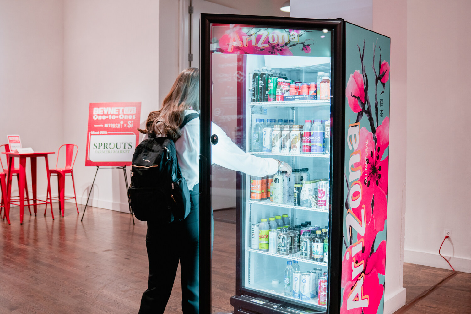 A woman with a backpack opens a glass-door fridge wrapped in Arizona graphics with cherry blossoms, beside a Sprouts One-to-Ones sign.
