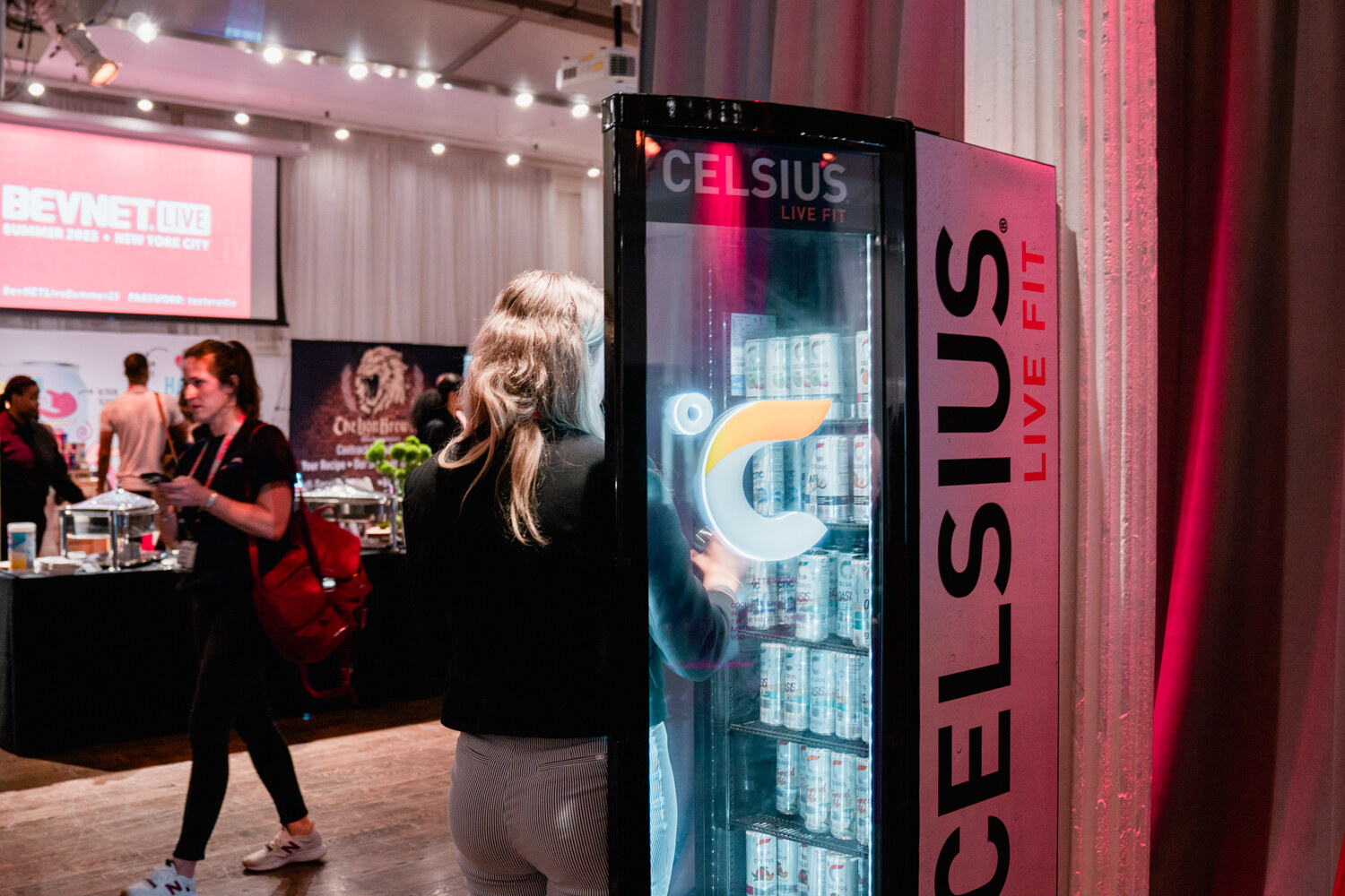 Attendees walk past a glass-door Celsius Live Fit fridge in a loft room with catering tables in the background.