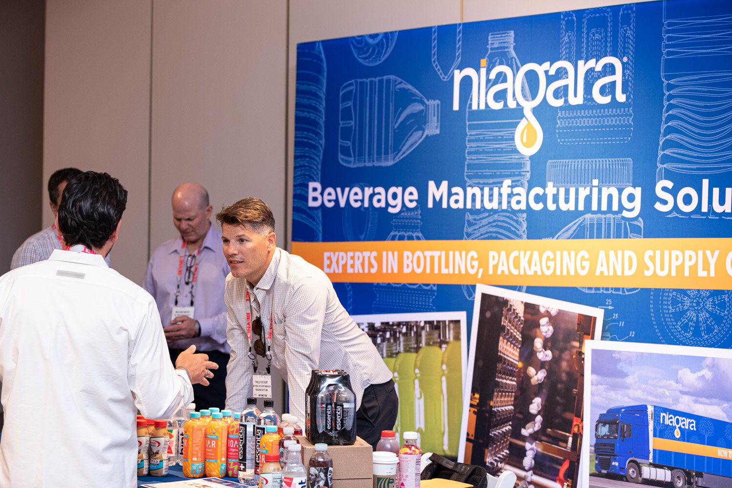 A man in a white shirt leans on a table of bottles in front of a blue Niagara banner reading Beverage Manufacturing Solutions.