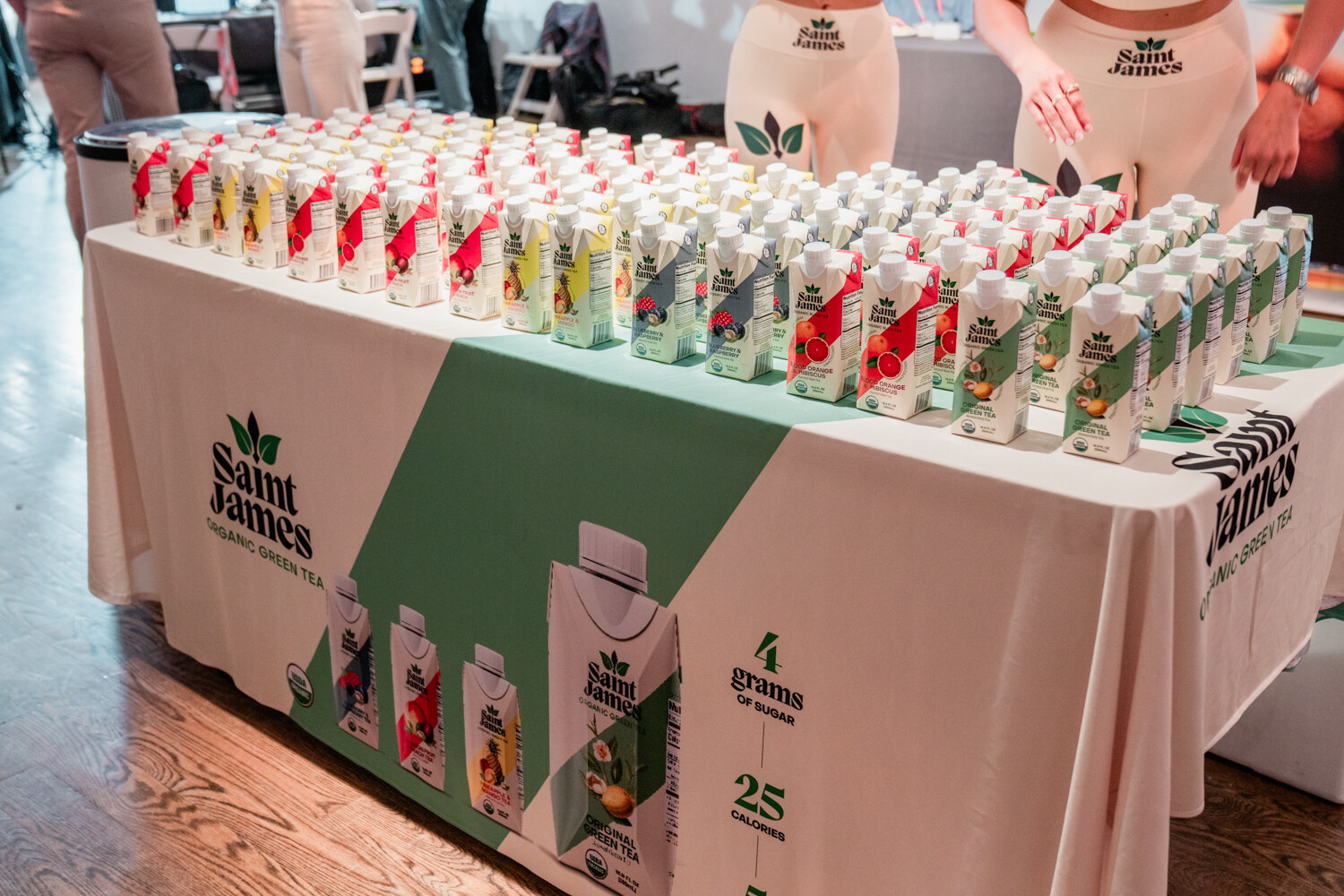 A long table covered with a white and green Saint James cloth holds rows of small iced tea cartons, with staff in Saint James shirts behind it.