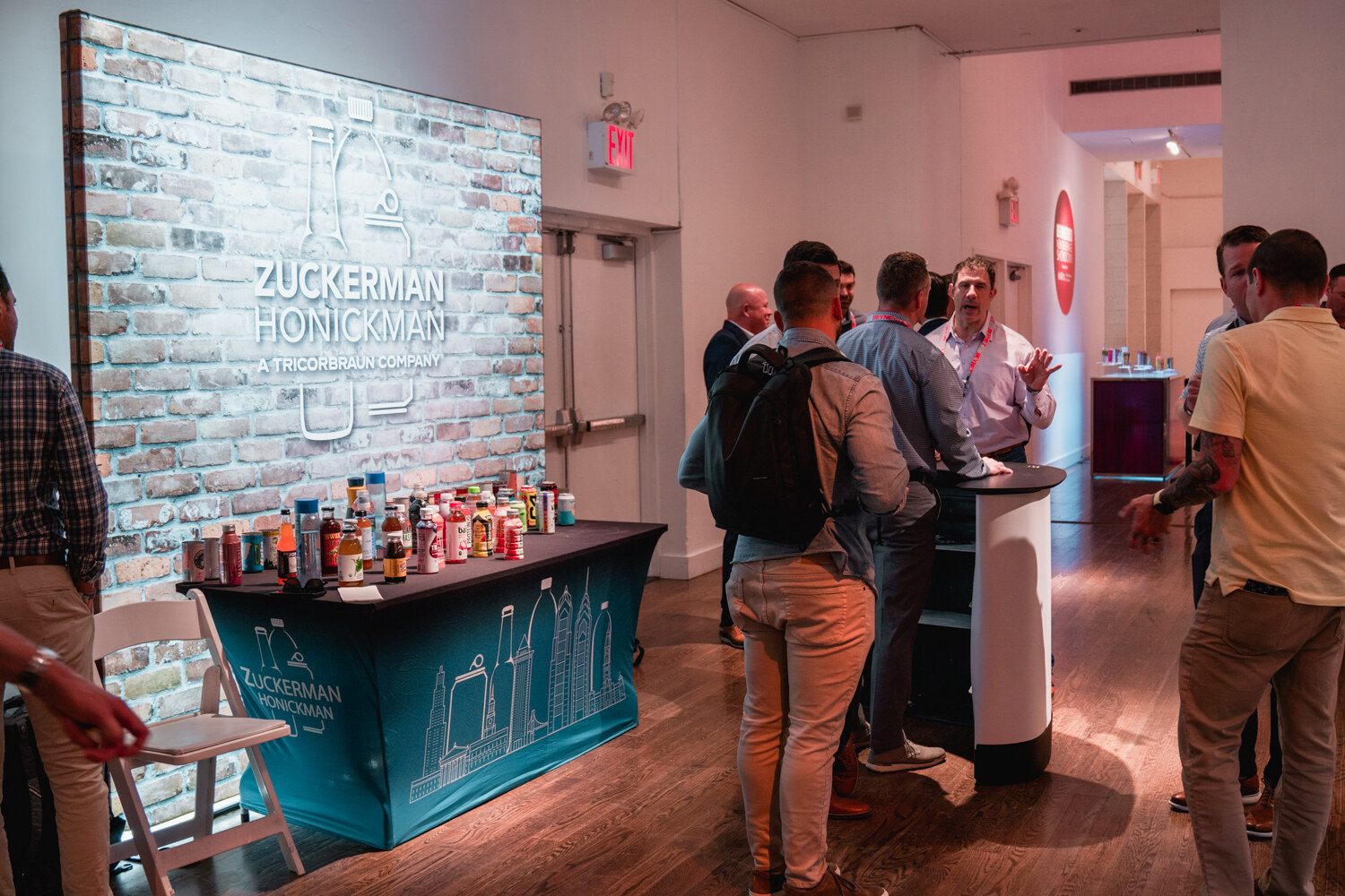 A teal draped counter displays bottles and jars in front of a brick-pattern wall with a Zuckerman Honickman logo while attendees stand around.