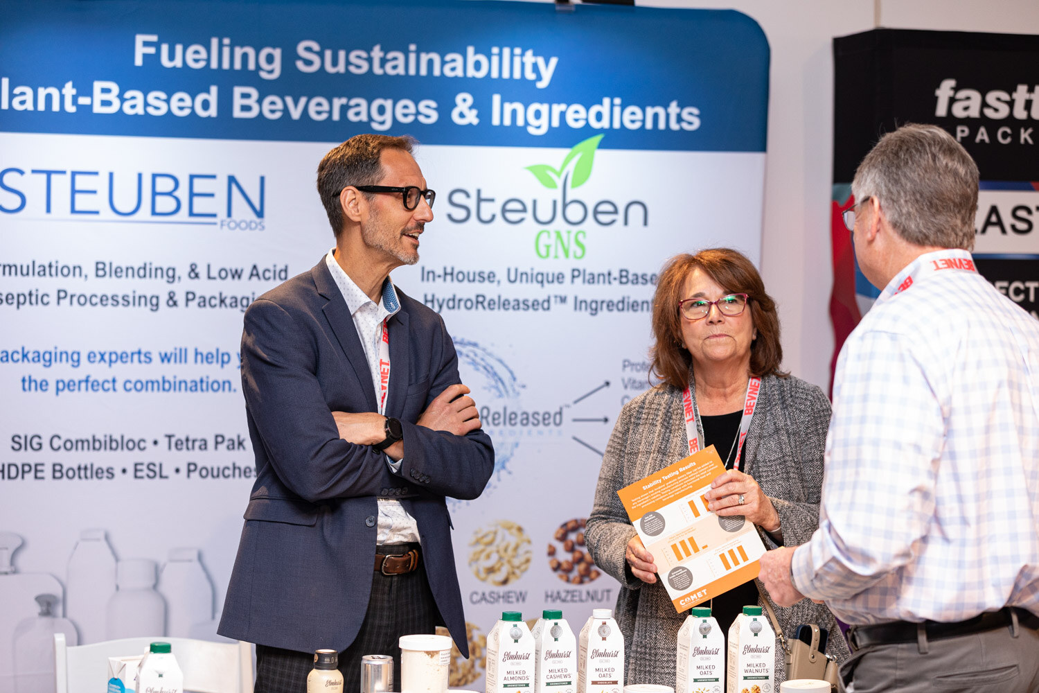 A man in a blazer with crossed arms, a woman holding a brochure, and a man in a checked shirt stand in front of a Steuben Foods banner about plant-based beverages and ingredients.