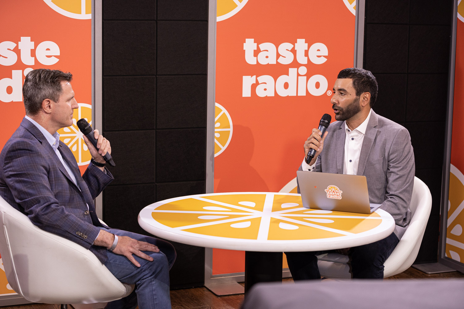 A man in a gray blazer holding a laptop-side microphone speaks with a man in a navy blazer at a round orange-slice table in front of a Taste Radio backdrop.
