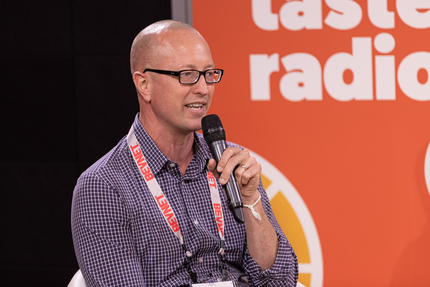 A bald man in glasses with a checked shirt and red lanyard speaks into a microphone in front of an orange Taste Radio backdrop.