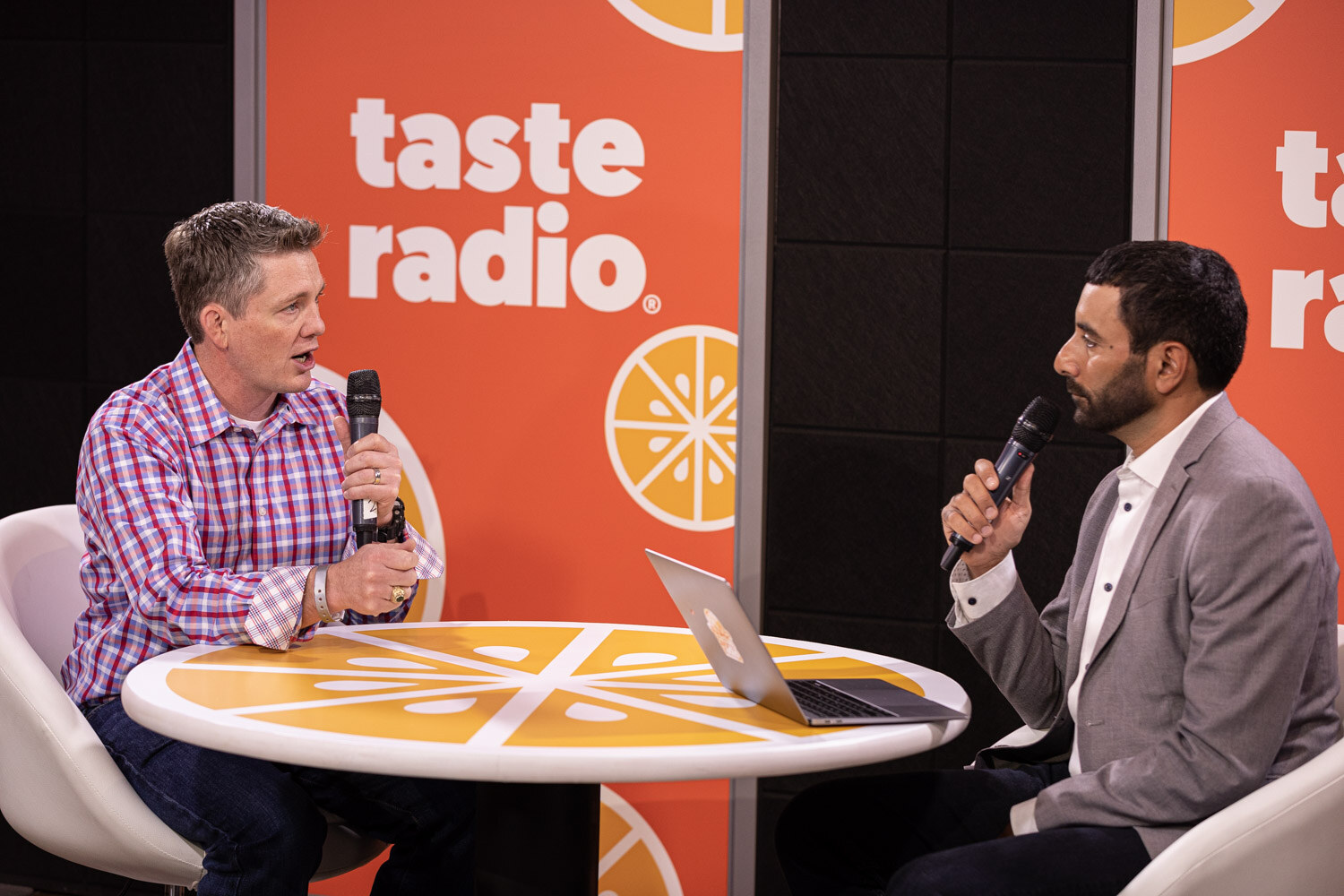 A man in a pink plaid shirt and a man in a gray blazer sit at a round orange table with a laptop, both holding microphones.