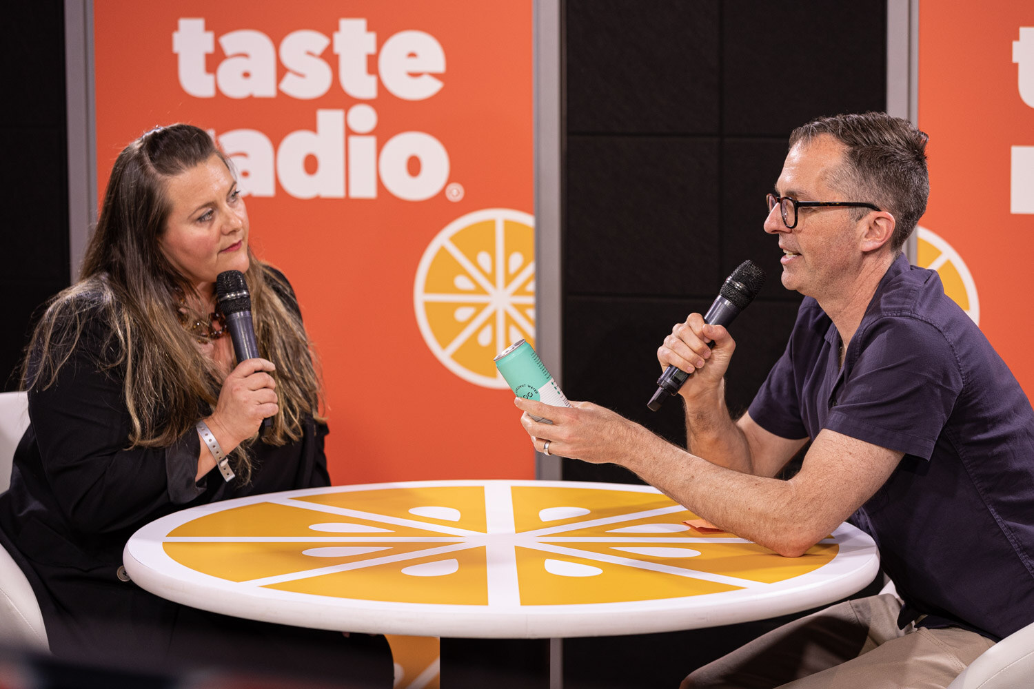 A woman in black and a man in a navy shirt with glasses sit across a round orange table, with the man holding out a small teal can.