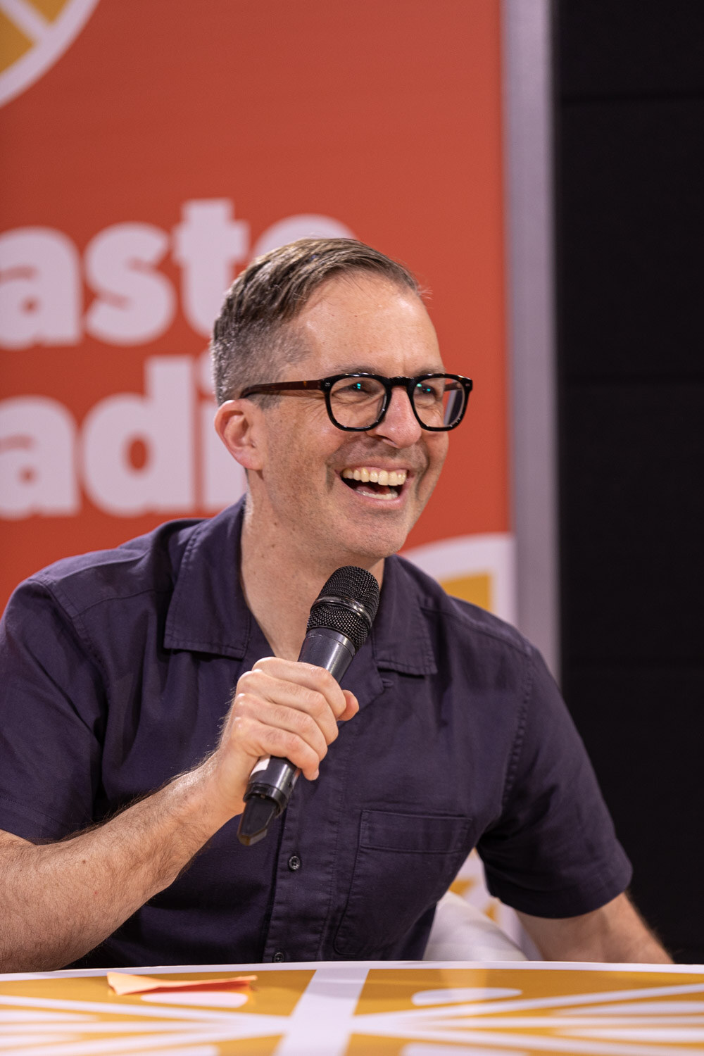 A smiling man with glasses and a dark navy short-sleeve shirt holds a microphone in front of an orange Taste Radio backdrop.