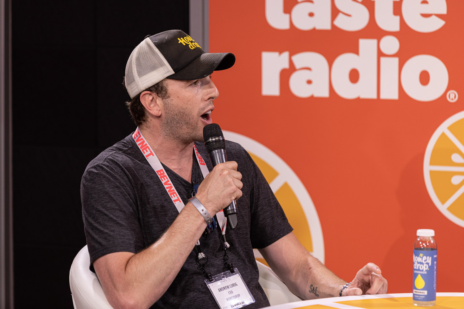 A man in a mesh-back trucker cap and dark T-shirt with a lanyard speaks into a microphone in front of a Taste Radio backdrop with a juice bottle beside him.