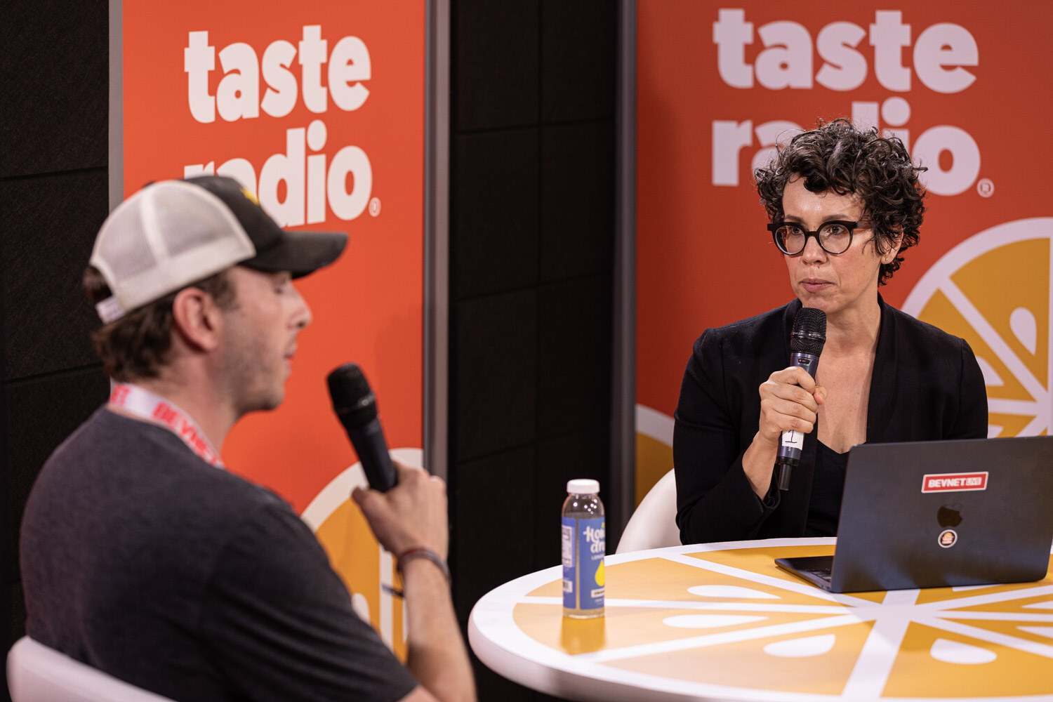 A man in a mesh-back cap faces a woman with curly hair and dark glasses who holds a microphone, with a laptop and a bottle on the orange table.