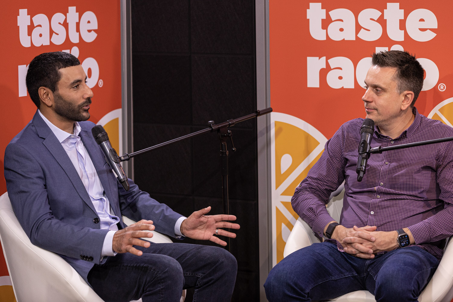 A man in a blue blazer and a man in a purple checked shirt sit in white chairs in front of an orange Taste Radio backdrop with boom microphones around them.