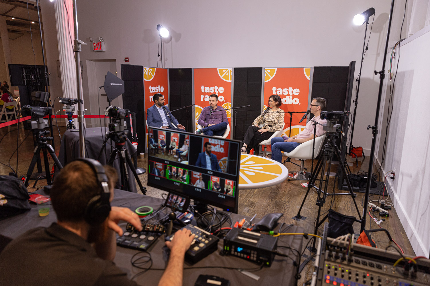 View from behind studio cameras and a monitor wall toward the orange Taste Radio set where four people sit under lights.