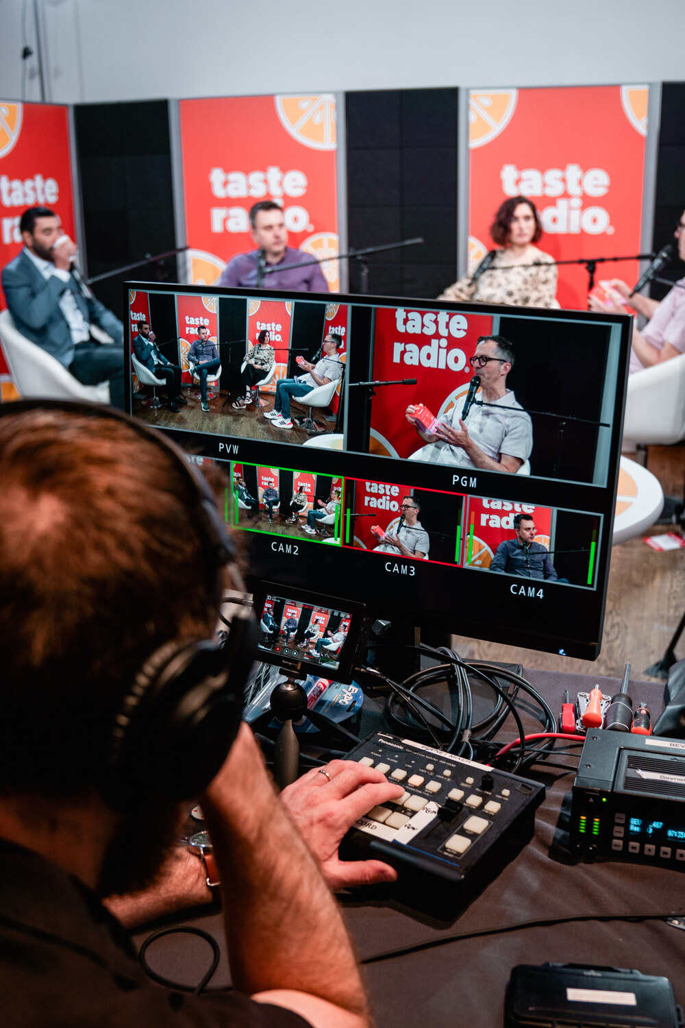 A person wearing headphones sits at a video switcher in front of a monitor showing several camera views of the Taste Radio studio.