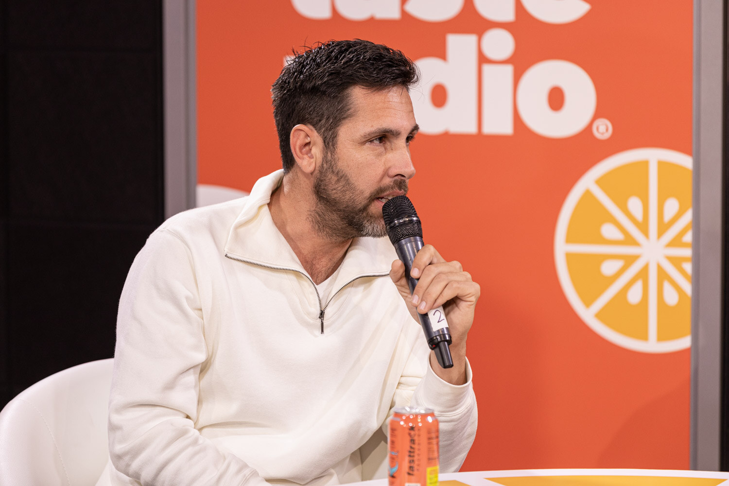 A man with dark hair and a beard in a cream quarter-zip sweater holds a microphone with an orange can on the table in front of him.