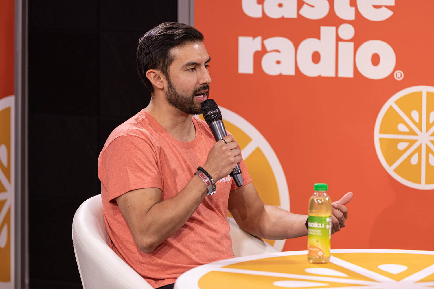 A man with dark hair and a beard in a coral T-shirt holds a microphone with a green juice bottle on the table in front of an orange backdrop.