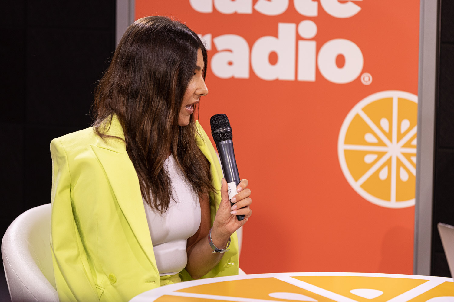 A woman with long dark hair in a lime-yellow blazer and white top holds a microphone in front of an orange Taste Radio backdrop.