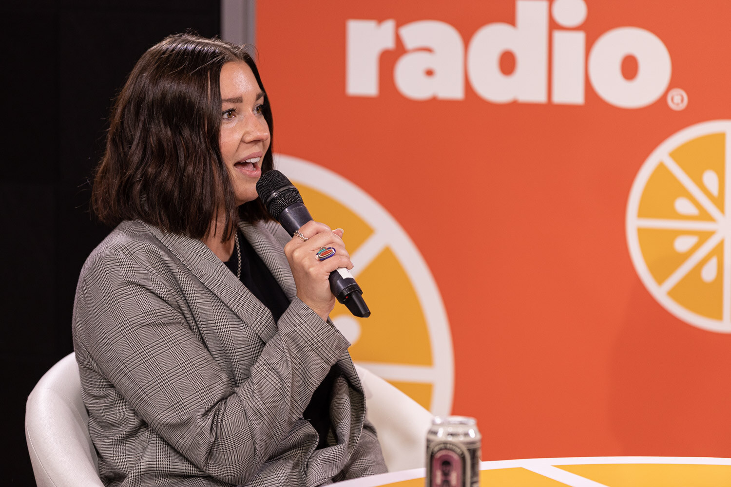 A woman with a dark bob in a gray blazer holds a microphone with a can on the table in front of an orange Taste Radio backdrop.