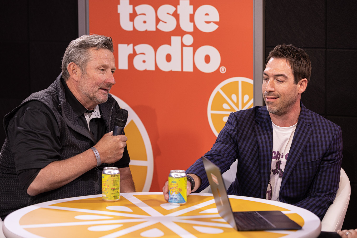 A gray-haired man in a black vest holds a microphone across from a man in a patterned navy blazer, with yellow cans and a laptop on the orange table.