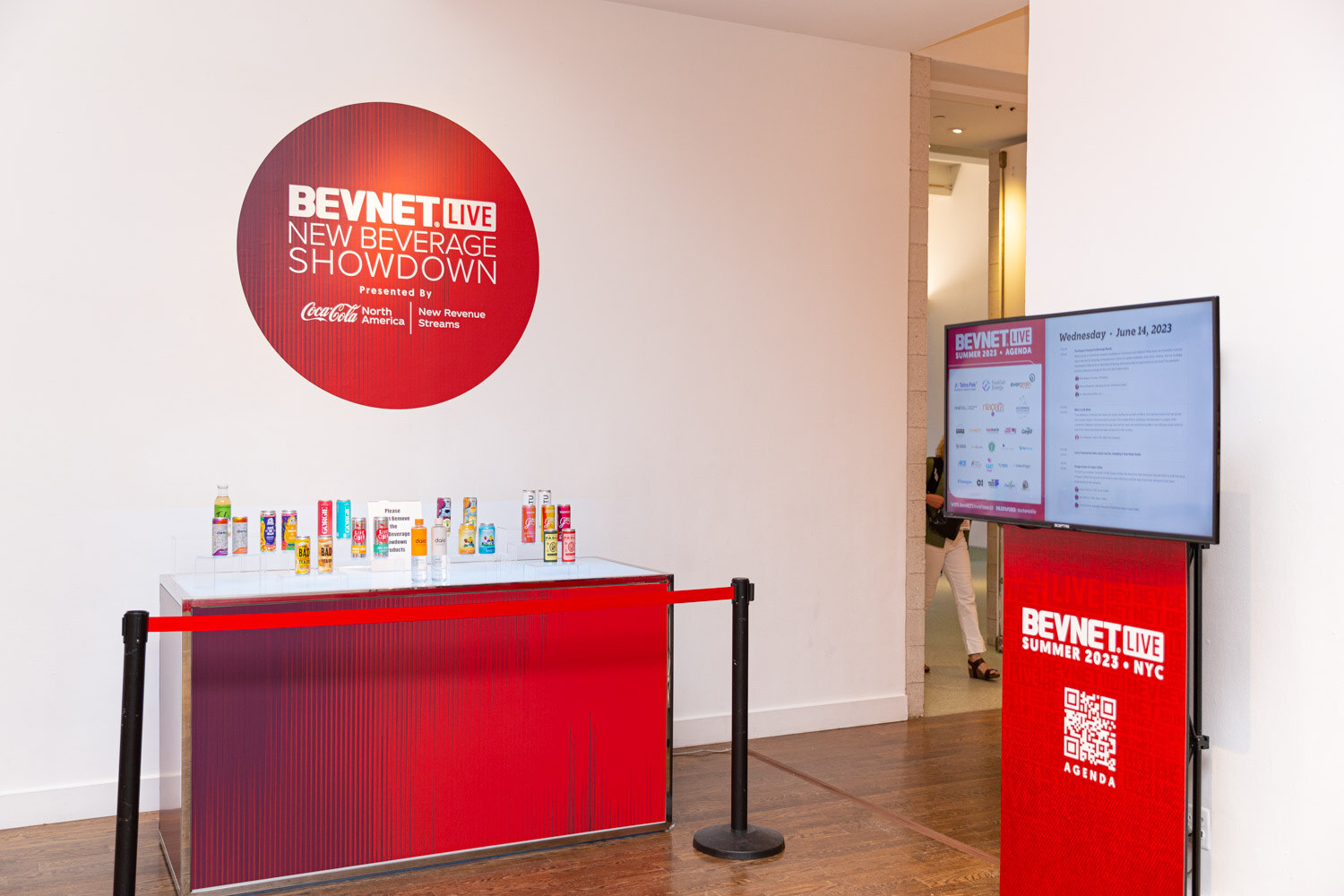 A red table with a row of colorful drink cans and bottles sits below a round red BevNET Live New Beverage Showdown logo presented by Coca-Cola North America New Revenue Streams.