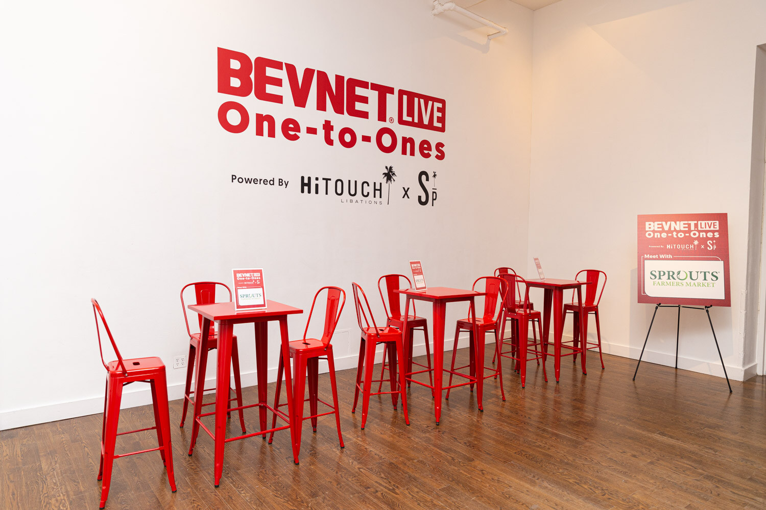 A white room with red high tables and stools, a BevNET Live One-to-Ones sign on the wall, and a Sprouts sign on an easel.