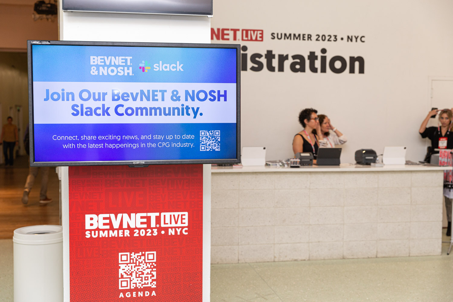 A monitor above a red pedestal with a QR code reads Join Our BevNET and Nosh Slack Community, with the registration desk and staff behind it.