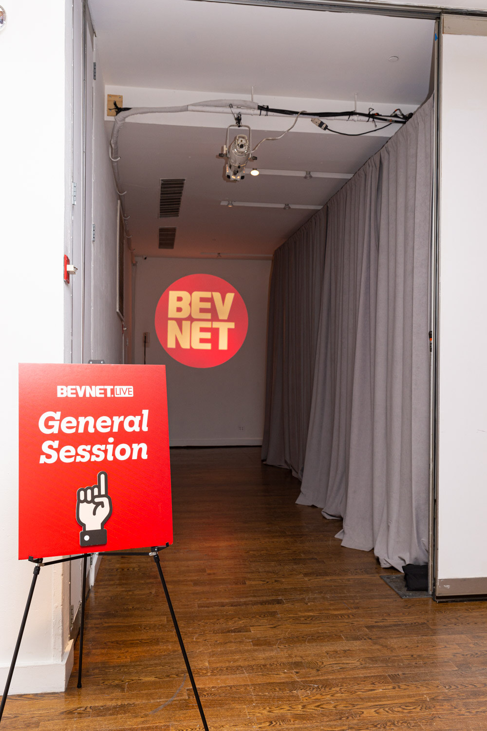 A red easel sign reading General Session stands in a narrow hallway lined with curtains with a round BevNET logo on the wall.