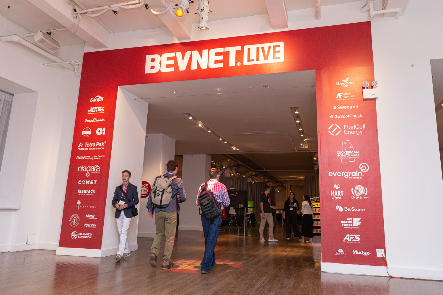 A red arch reading BevNET Live and covered in sponsor logos frames a wide hallway as attendees with backpacks walk through.