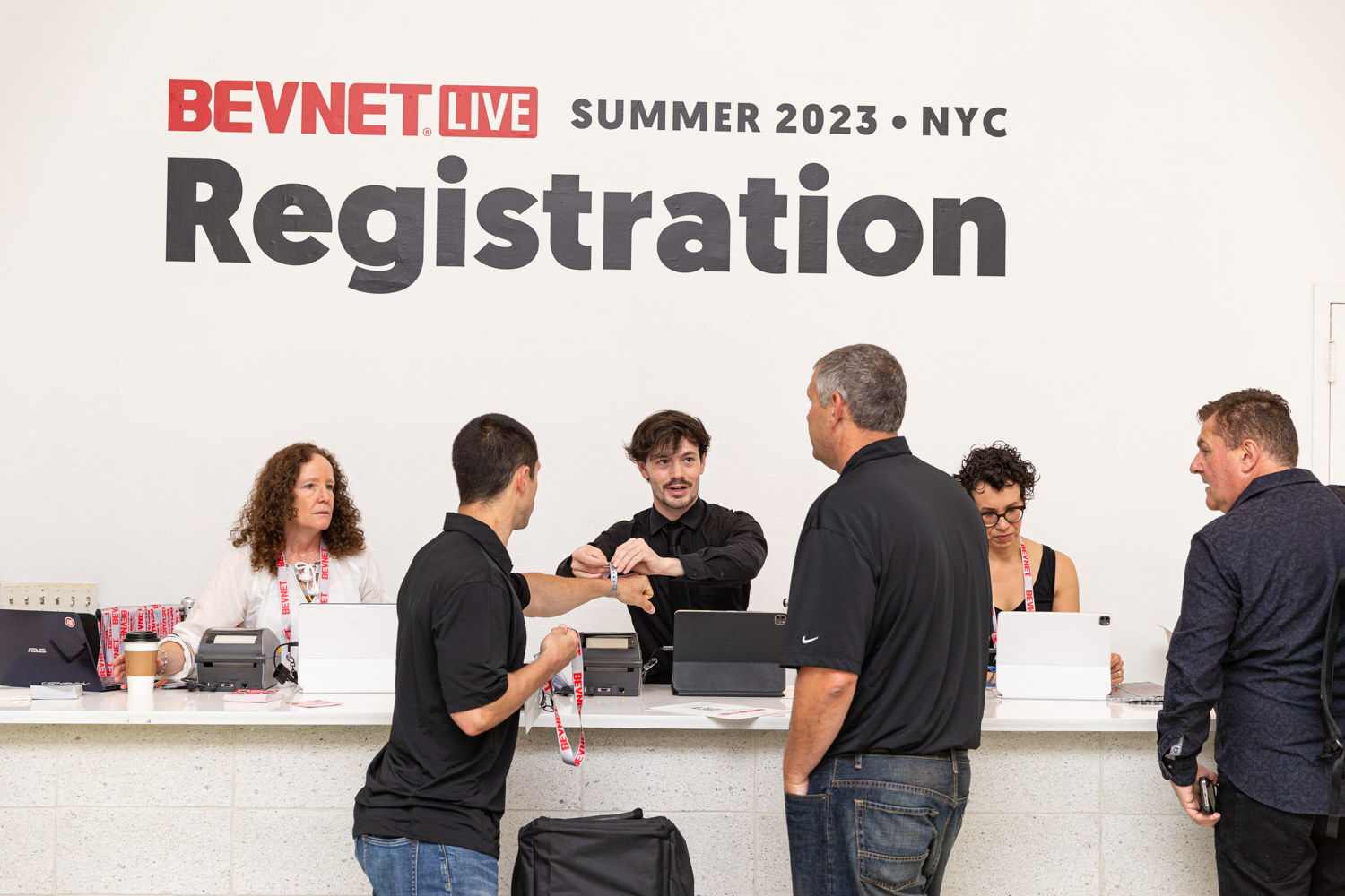 Several staff in black shirts and attendees stand at a white counter with laptops beneath a wall sign reading BevNET Live Summer 2023 NYC Registration.