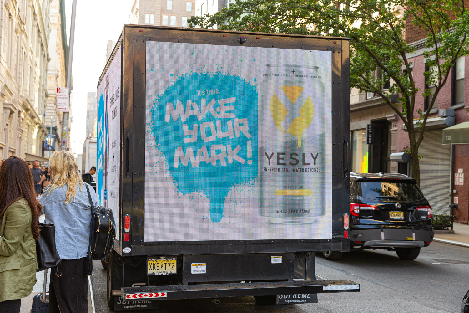 The side of a truck with a large blue graphic reading Make your mark and a Yesly can, parked on a city street with pedestrians nearby.