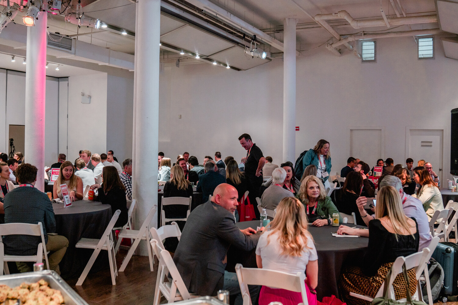 Attendees sit at round tables with black tablecloths in a loft room with white columns, with a tray of pastries at the lower left.