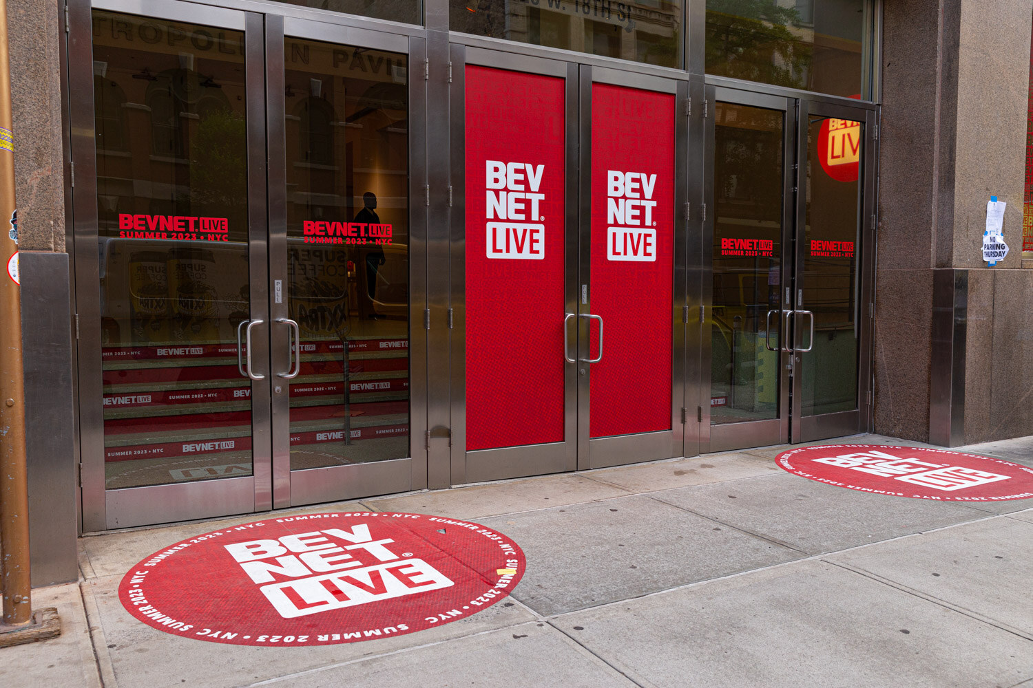 Glass and steel double doors wrapped in red BevNET Live graphics with round red BevNET Live decals on the sidewalk in front.