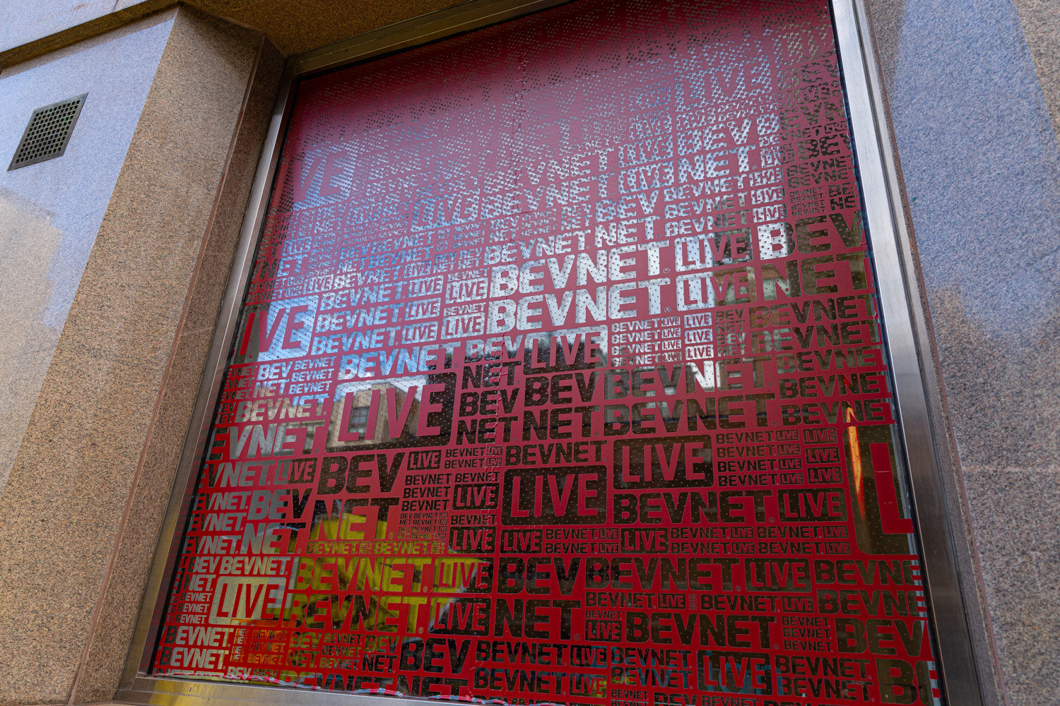A large ground-floor window covered with a red film printed with a repeating pattern of BevNET and Live logos, set in a stone building facade.