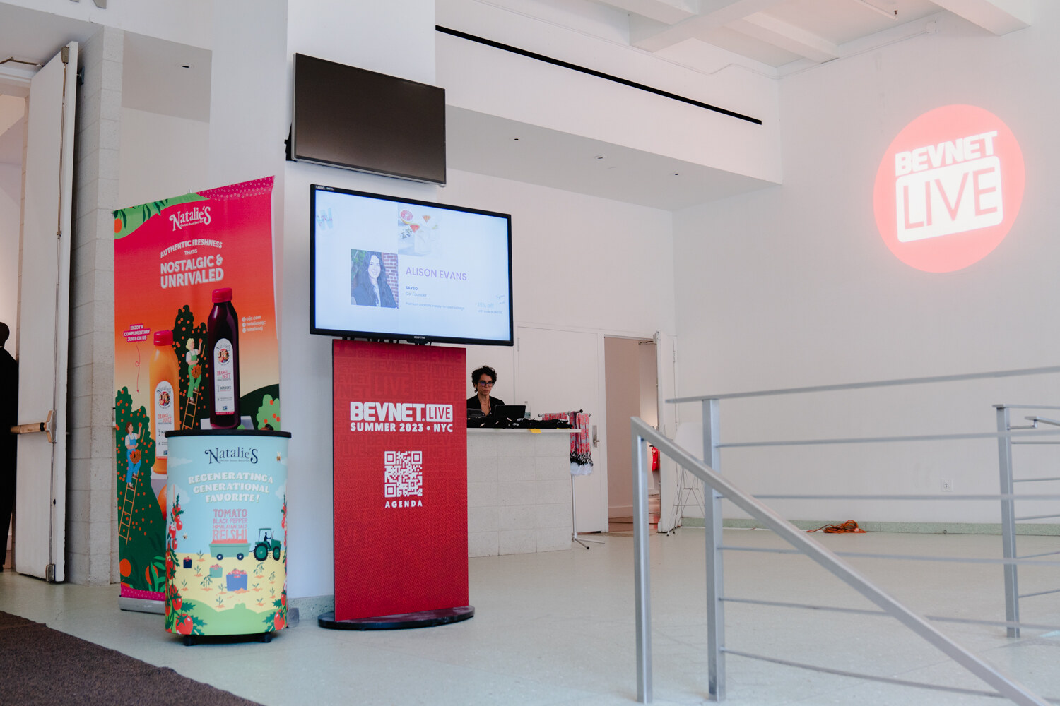 A colorful Natalie's juice display and a red BevNET Live pedestal with a screen stand near a registration counter and a lit round BevNET Live projection.