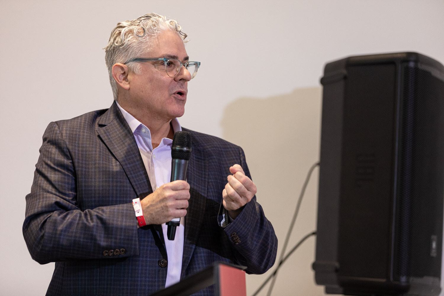 Presenter in a plaid blazer and glasses speaks beside a loudspeaker at Beverage School.