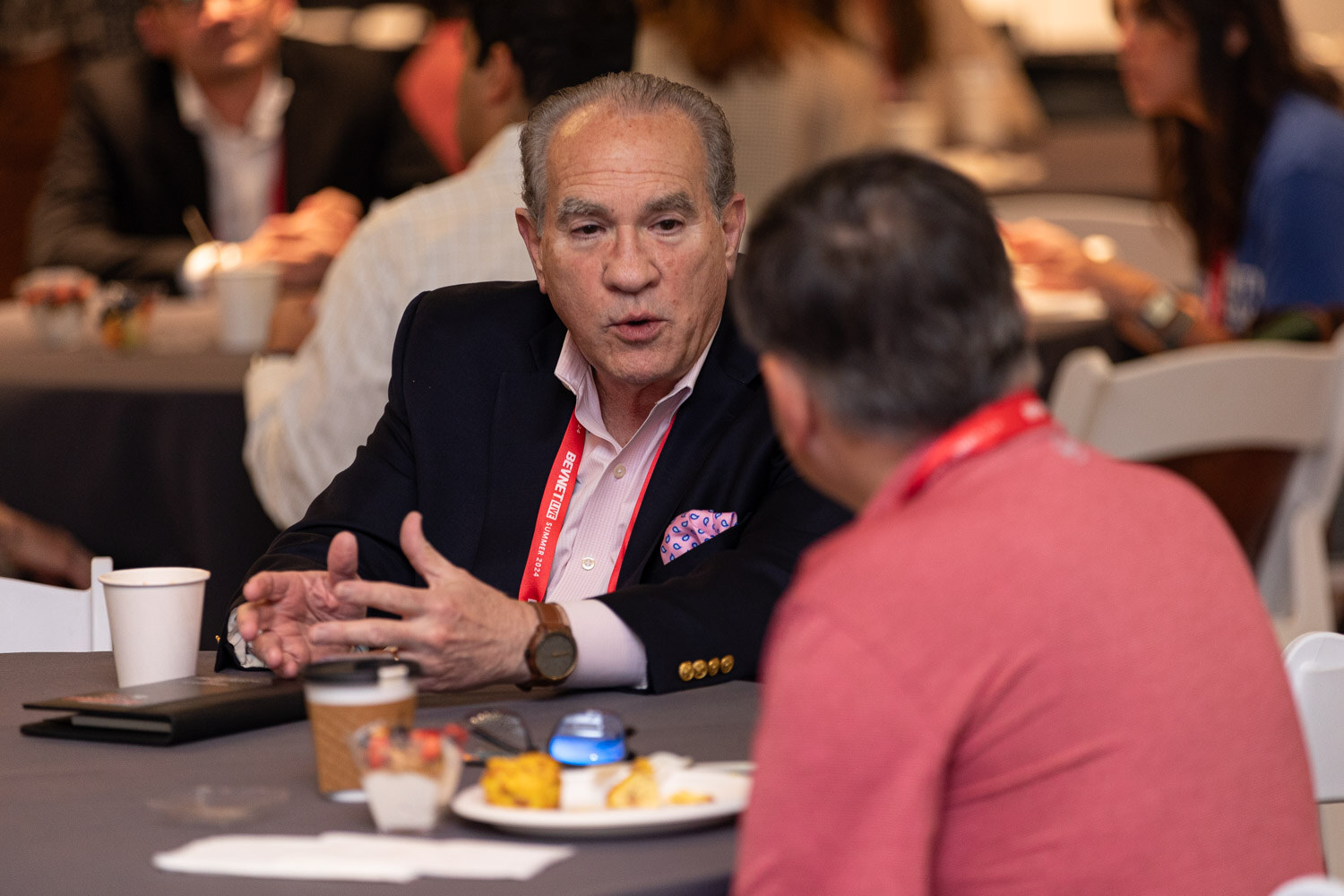 Attendee in a navy blazer talks with a seated colleague at a networking table.