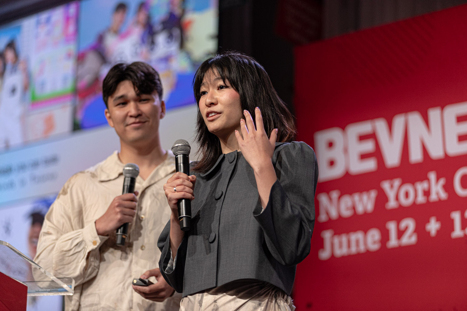 Two finalists hold microphones at the podium in front of a collage slide.