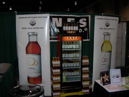 Booth with a lit refrigerated cooler of bottled drinks between two tall Jones Organics banners showing bottles, with product stacked beside the cooler and a table of signs at the right.
