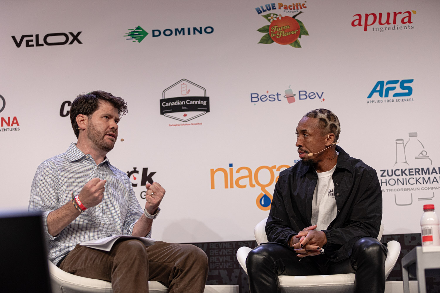 Moderator in a checked shirt and a guest in a black jacket sit on stage during Sports Marketing.
