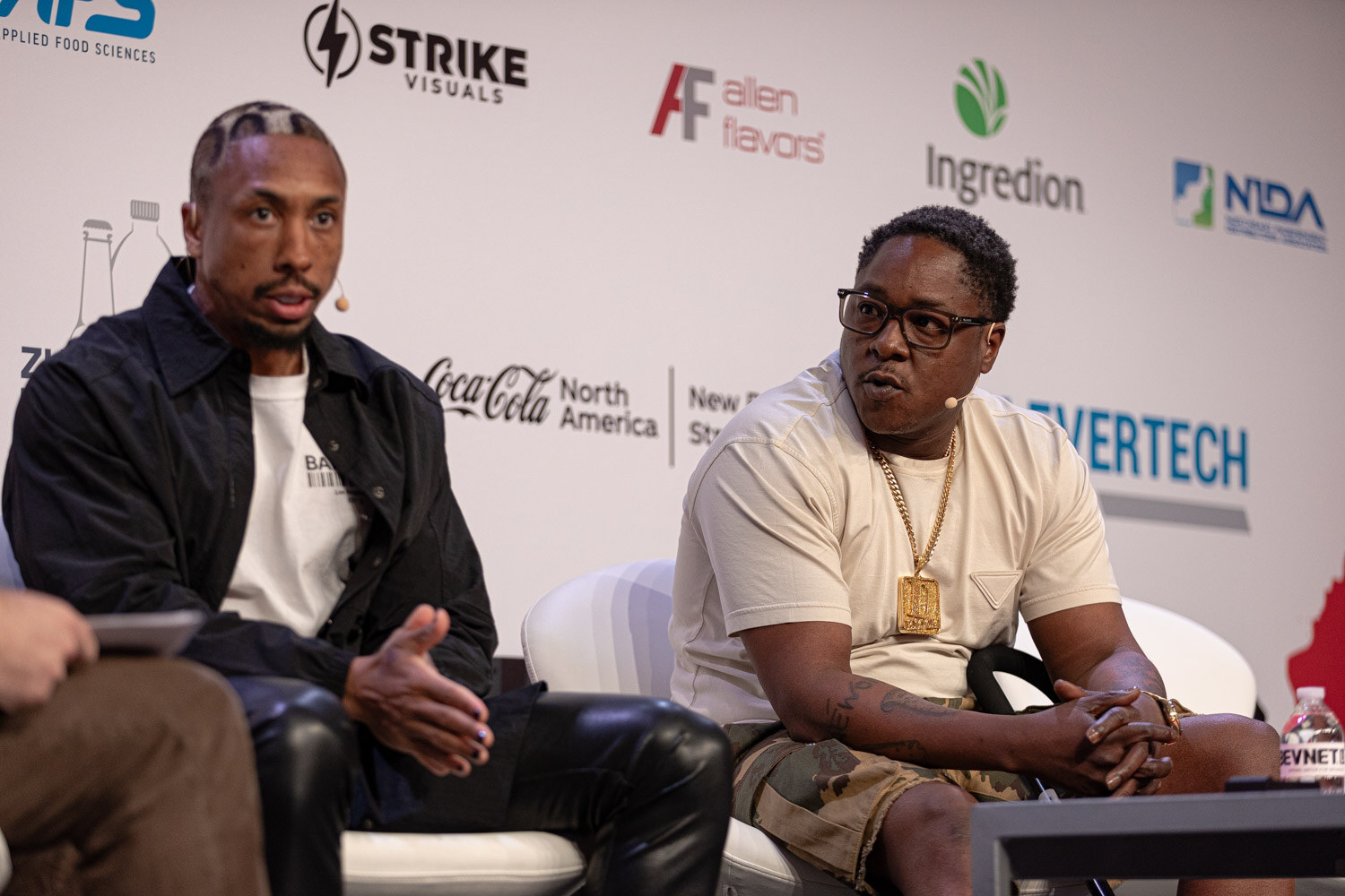 Two guests, one in a black jacket and one in a cream t-shirt with a gold pendant, sit side by side.
