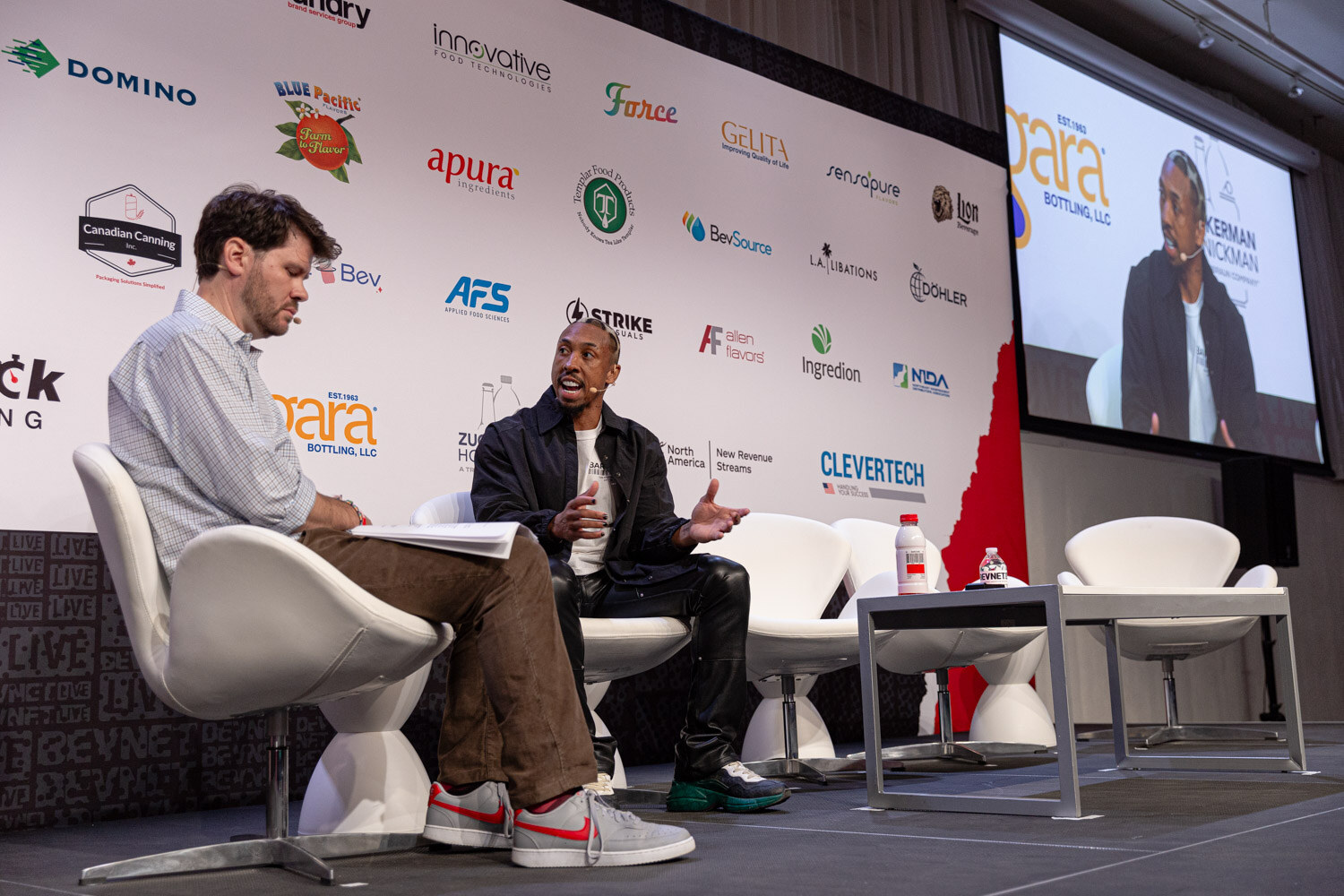 Wide view of the moderator and guest on stage next to a screen during Sports Marketing.