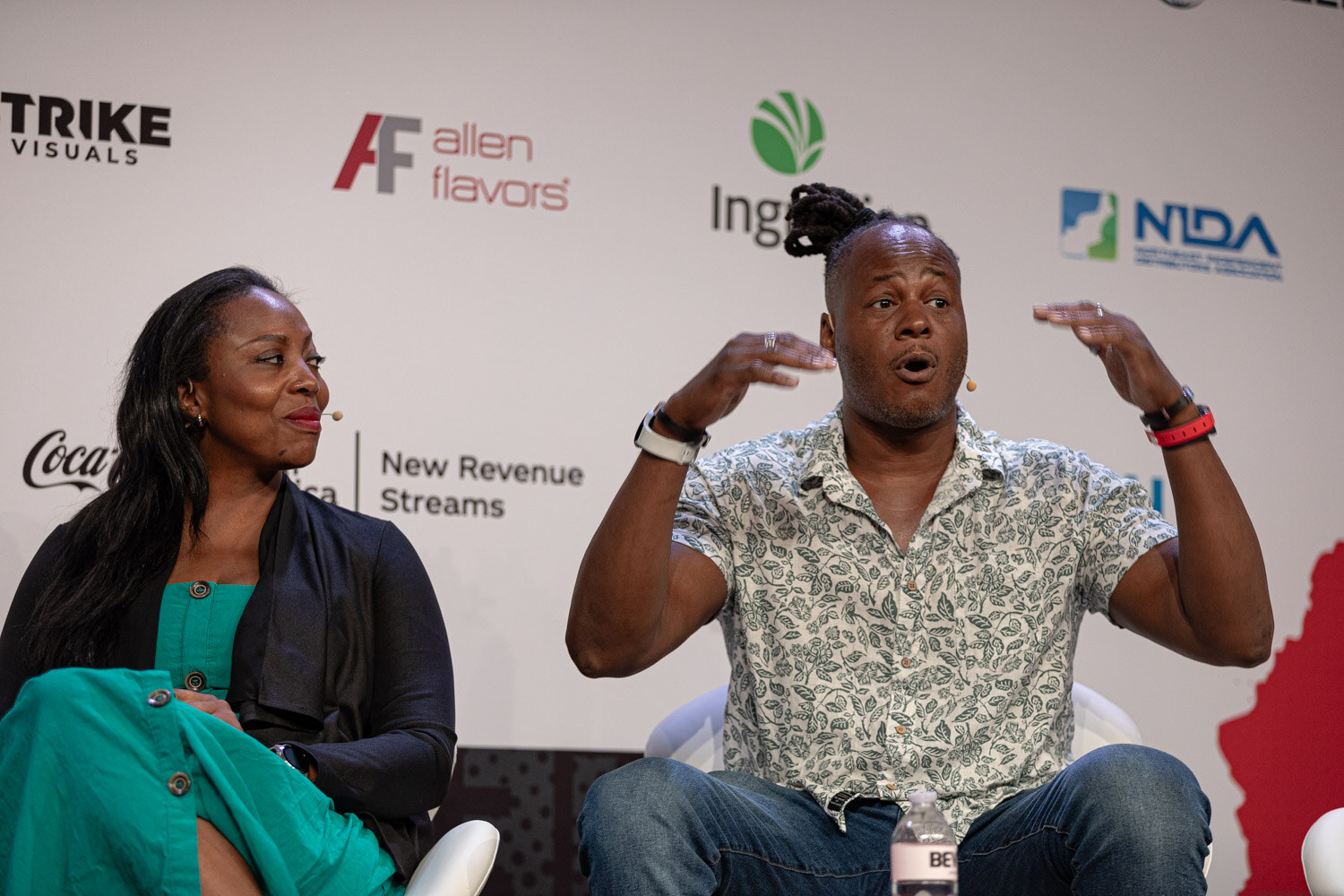 Panelist in a patterned shirt speaks while a panelist in a teal outfit listens.