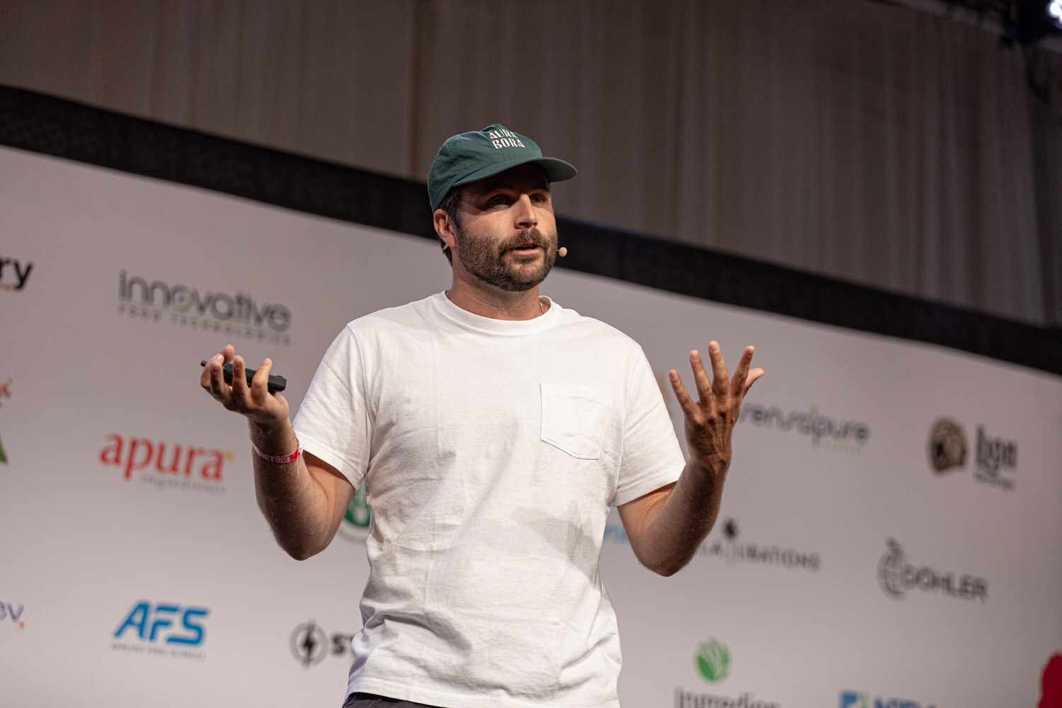 Speaker in a white t-shirt and green cap gestures during The Fundraising Journey.