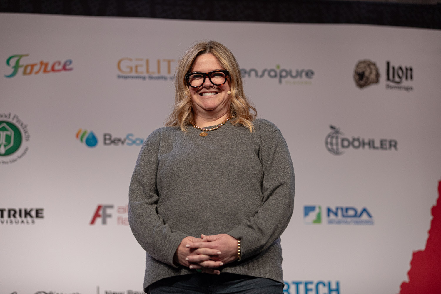 Co-presenter in a gray sweater and black glasses stands smiling on stage.