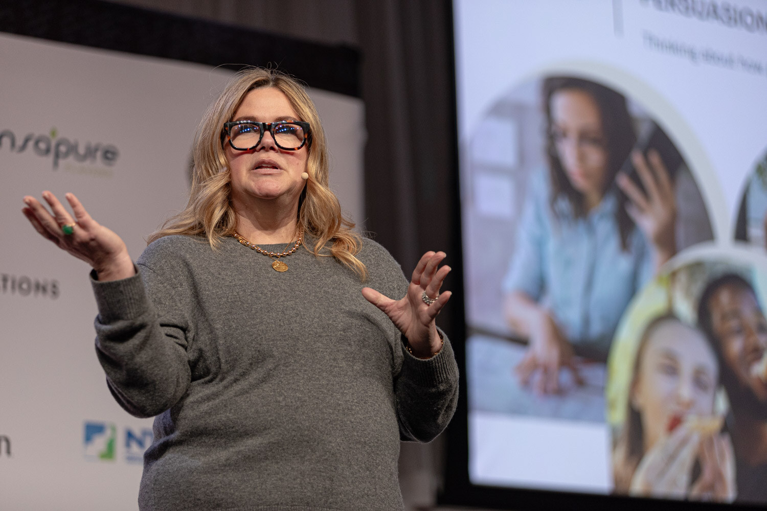 Co-presenter in a gray sweater explains a point with open hands.