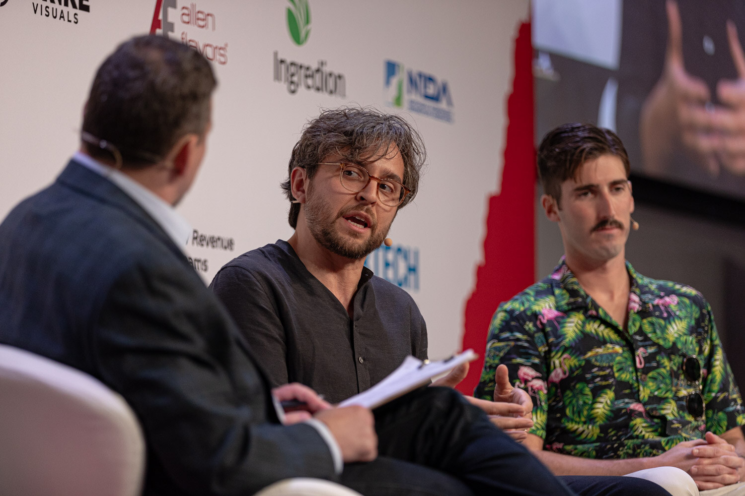 Panelist in a dark linen shirt speaks while the moderator takes notes.