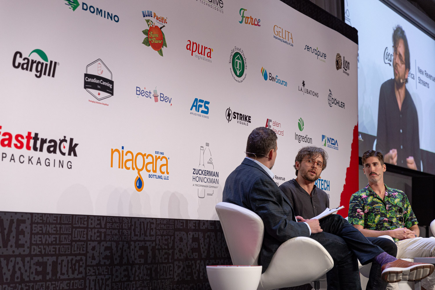 Moderator and two panelists seated on stage in front of the sponsor wall.