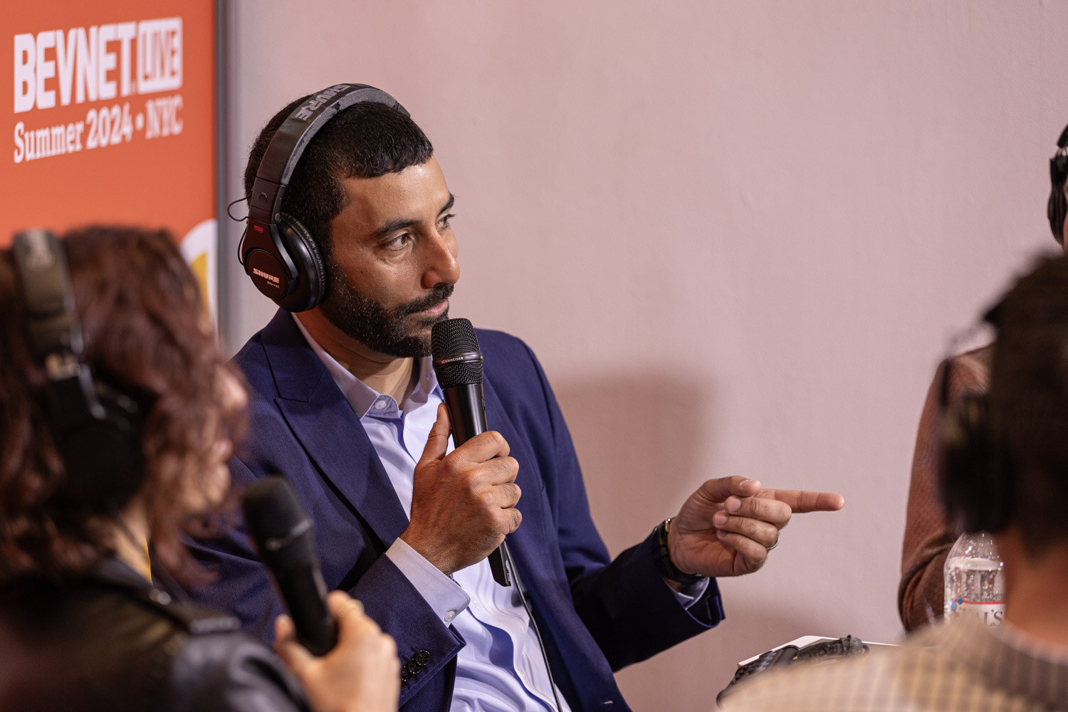 Host in a blue blazer points while speaking in headphones during the livestream.