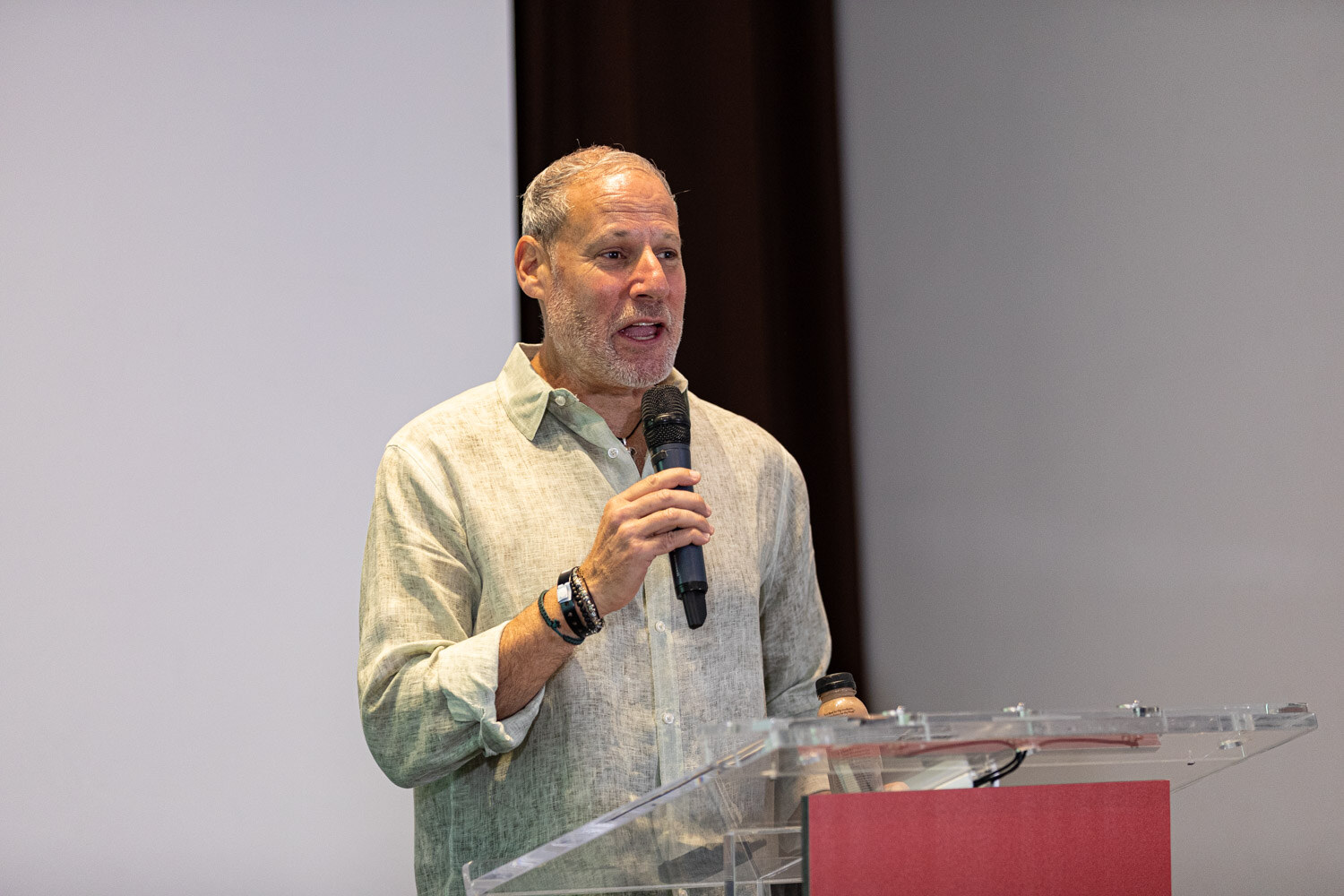 A gray-haired man in a green linen shirt speaks into a microphone at a clear podium.