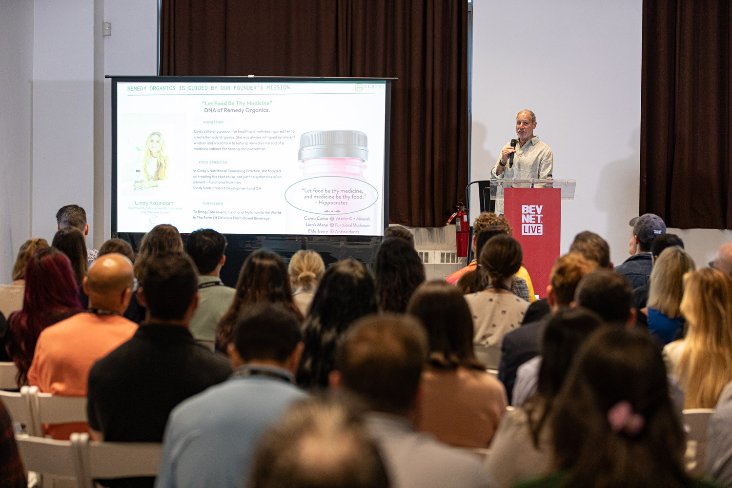 A crowded room of seated attendees facing a projector screen and a speaker at a BevNET Live podium.